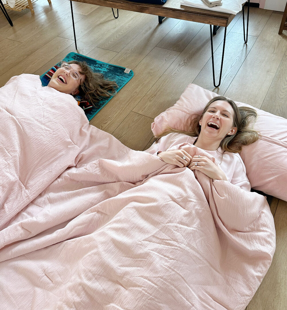 Two friends laughing under cozy pink blankets at a Tropical Refuge slumber party